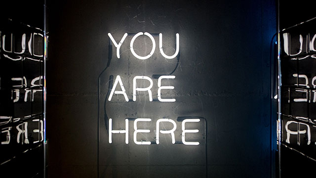 white neon sign reading You Are Here on black background
