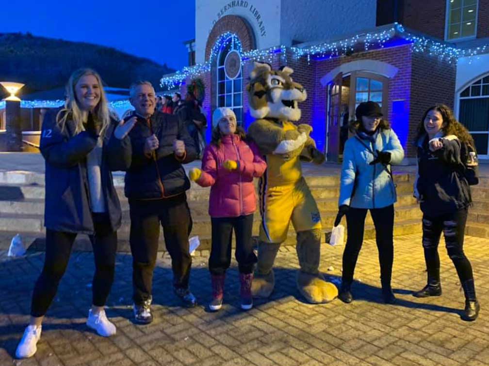 men and women dancing in winter coats outside with bobcat mascot in fron of building with blue holiday lights