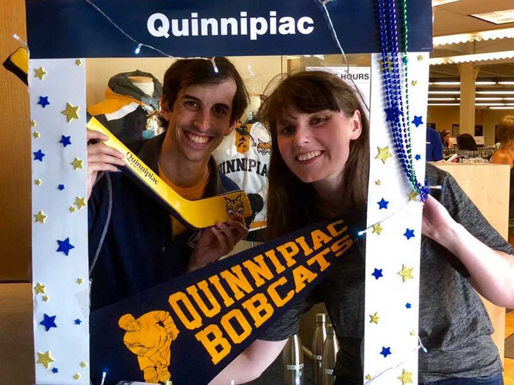 man and woman posing with blue and gold pennet and hockey stick in cardboard photo frame that says Quinnipiac